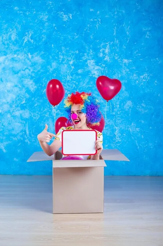 Dziewczyna klaun siedzi w kartonowym pudełku z balonami heliowymi i tablicą urodzinową