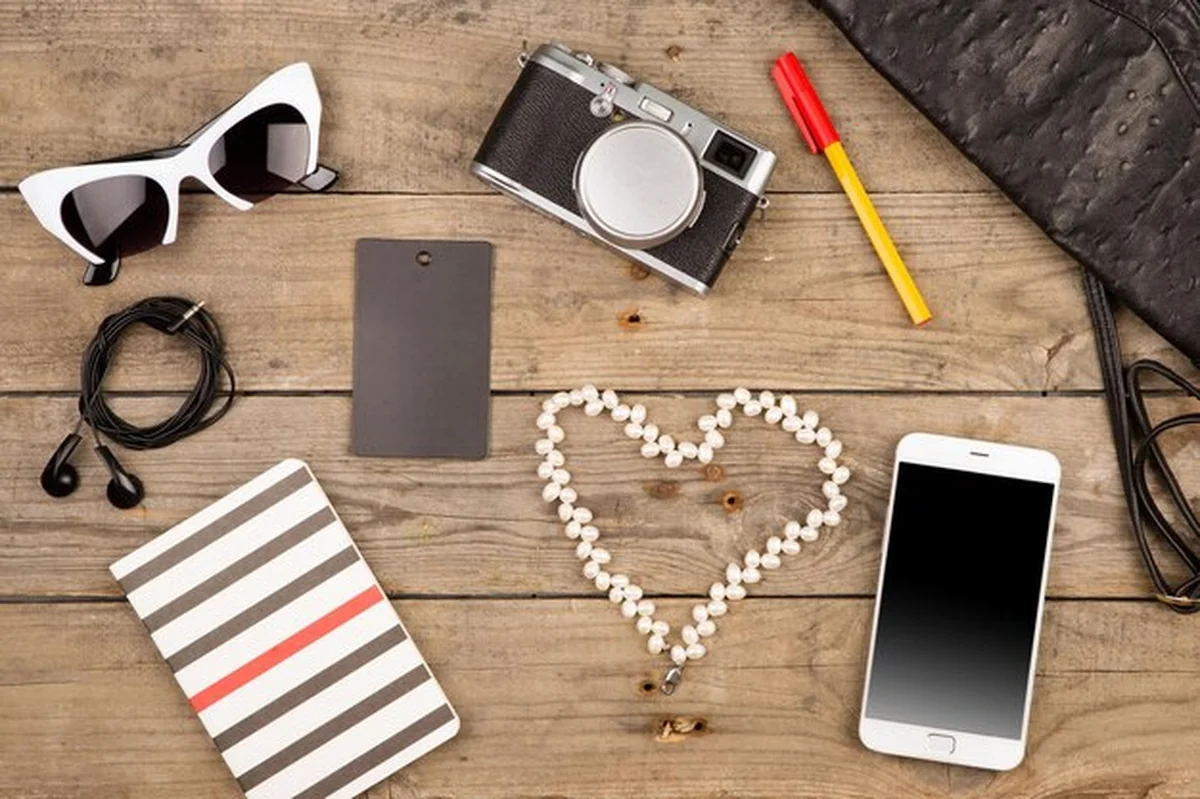 Zestaw damski na brązowym biurku: torba, smartfon, okulary przeciwsłoneczne, notatnik, słuchawki, aparat, gruszkowy tag