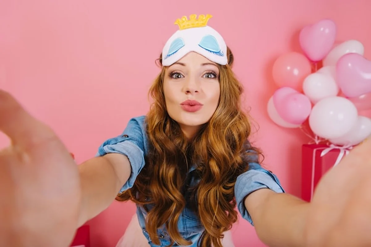 Młoda kobieta z balonami i prezentami, robiąca selfie w masce do spania przed urodzinową imprezą