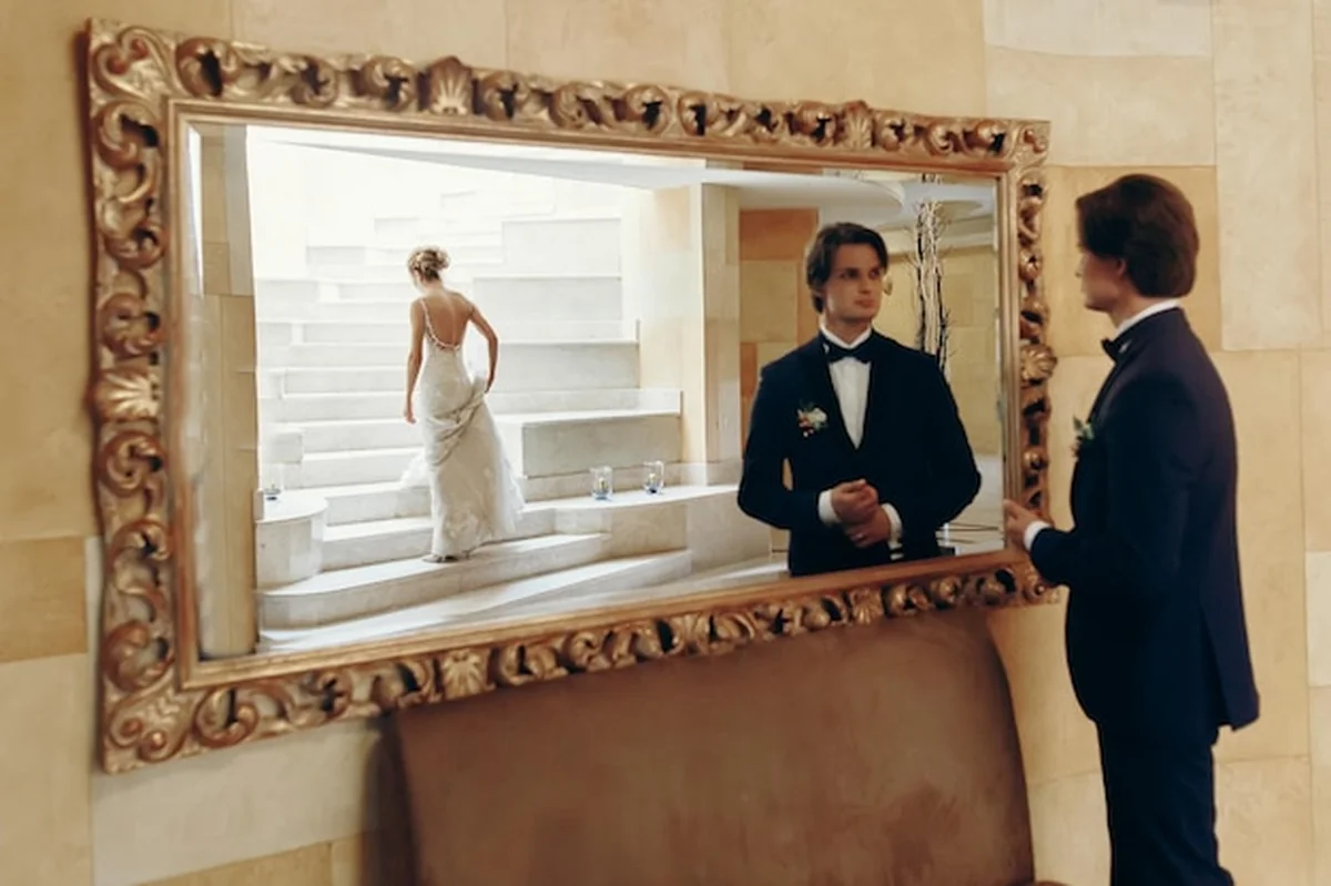 Młody pan w garniturze patrzy w lustro, obok blondynka w sukni ślubnej na białych schodach