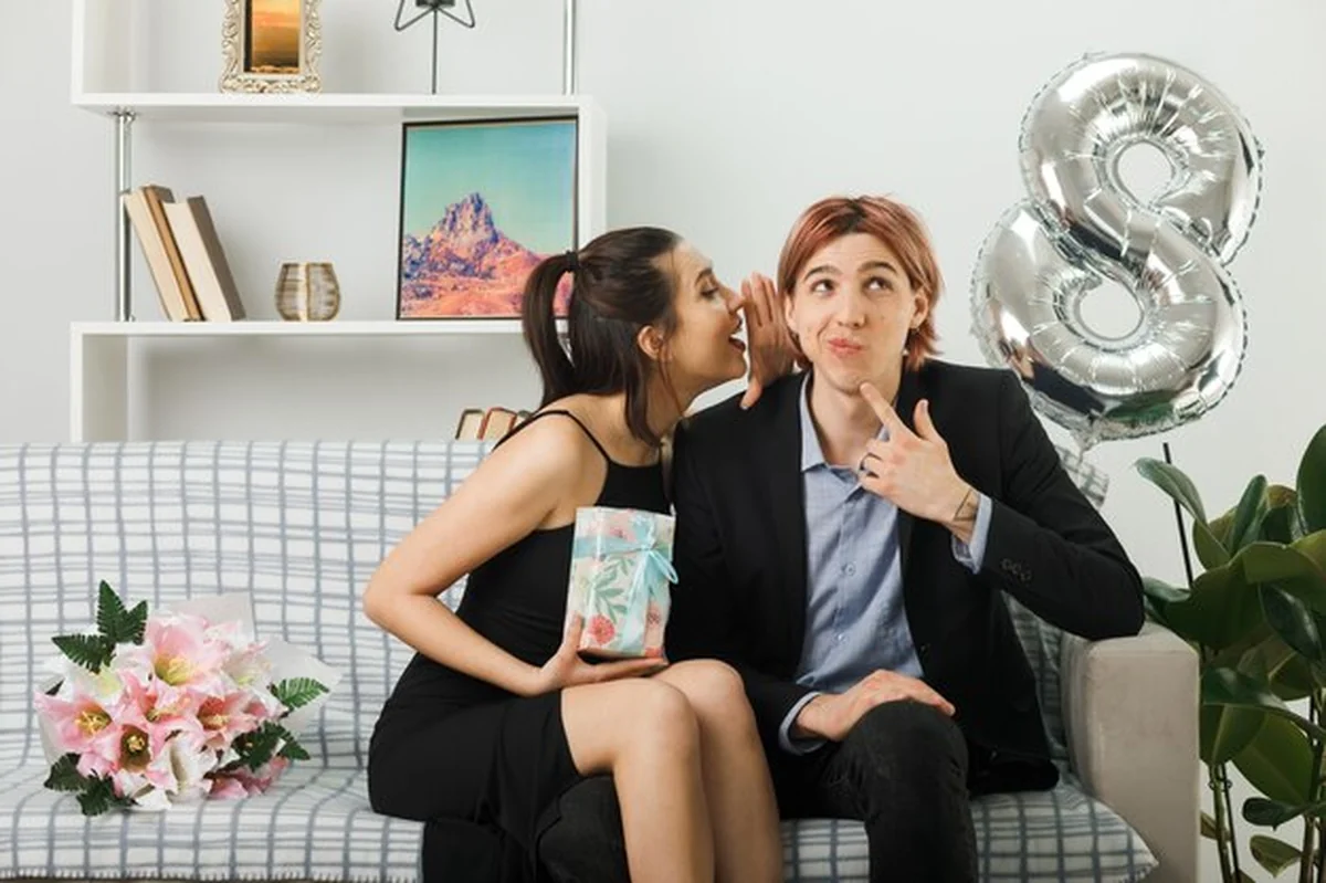 Młoda para w salonie: kobieta szepcze do mężczyzny siedzącego na kanapie