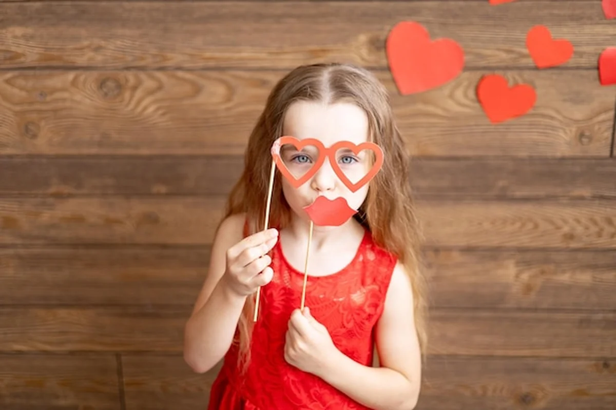 Mała dziewczynka w czerwonej sukience trzyma maskę okulary w kształcie serc na patyku na drewnianym tle z czerwonymi sercami