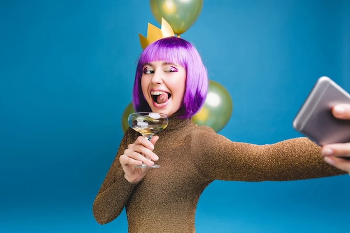 Młoda kobieta z fioletową fryzurą robi selfie z balonami, szampanem i pokazem języka na imprezie